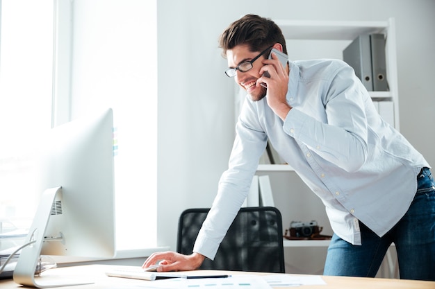 Feliz joven empresario de gafas trabajando con la computadora y hablando por teléfono celular en el lugar de trabajo