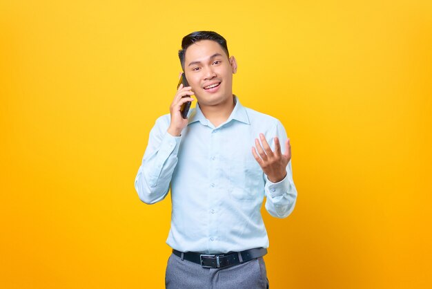 Feliz joven empresario guapo hablando por teléfono inteligente y mano levantada sobre fondo amarillo