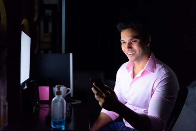 Feliz joven empresario indio usando el teléfono mientras trabaja horas extras en casa durante la cuarentena en la oscuridad