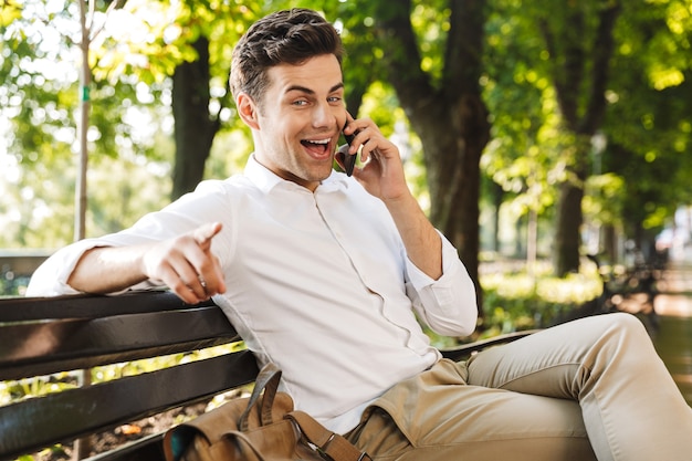 Feliz joven empresario sentado en un banco al aire libre, hablando por teléfono móvil