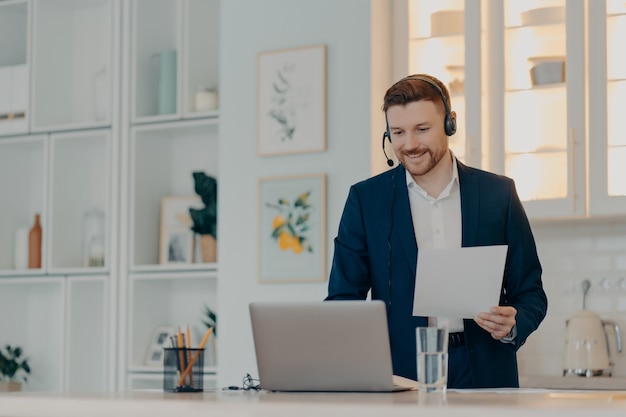 Feliz joven empresario con traje y auriculares trabajando de forma remota desde casa mientras usa la comunicación por video con sus colegas en la computadora portátil, sosteniendo el documento en la mano. Concepto de trabajo remoto