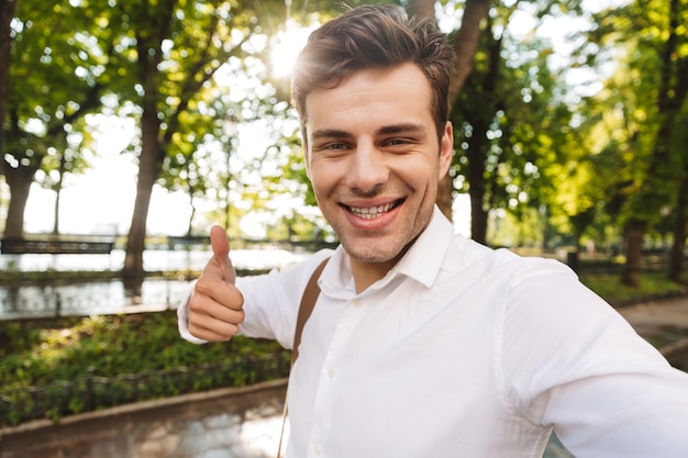 Feliz joven empresario vistiendo camiseta tomando un selfie mientras está de pie al aire libre en el parque de la ciudad, pulgares arriba