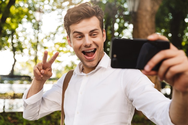 Feliz joven empresario vistiendo camiseta tomando un selfie mientras está de pie al aire libre en el parque de la ciudad, sosteniendo el teléfono móvil con la mano extendida