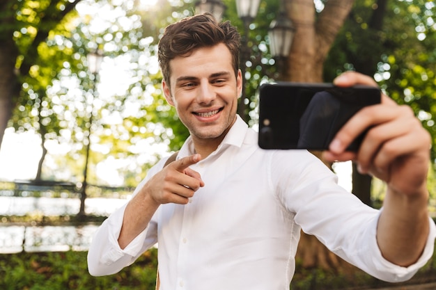 Feliz joven empresario vistiendo camiseta tomando un selfie mientras está de pie al aire libre en el parque de la ciudad, sosteniendo el teléfono móvil con la mano extendida