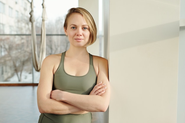 Feliz joven entrenador de yoga mosca en chándal gris manteniendo los brazos cruzados por el pecho mientras se inclina por la pared contra la hamaca en el gimnasio