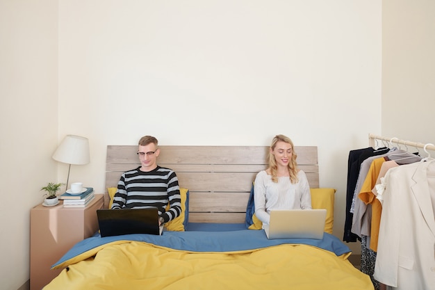 Feliz joven esposo y esposa sentados en la cama con computadoras portátiles, trabajando o jugando juegos en línea