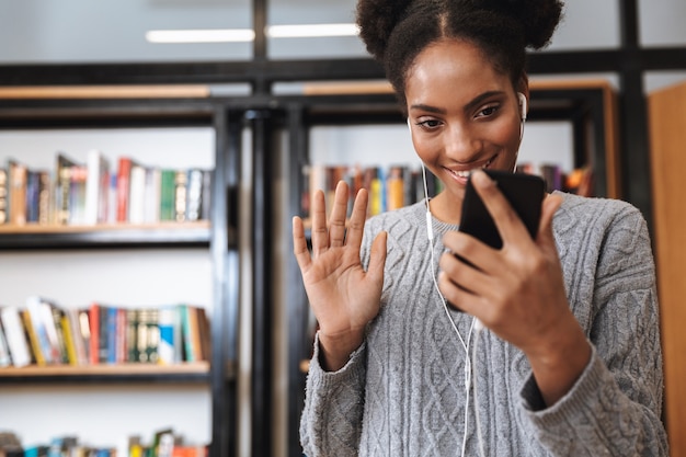 Feliz joven estudiante africana estudiando en la biblioteca, escuchando música con auriculares