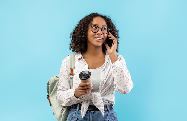 Feliz joven estudiante negra con mochila y café para llevar hablando en un teléfono inteligente en azul