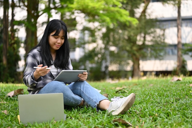 Feliz joven estudiante universitaria asiática usando su tableta digital mientras se relaja sentada en el césped