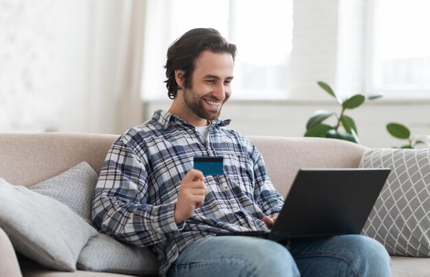 Feliz joven europeo adicto a las compras con rastrojo en camisa muestra tarjeta de crédito y mira la computadora portátil