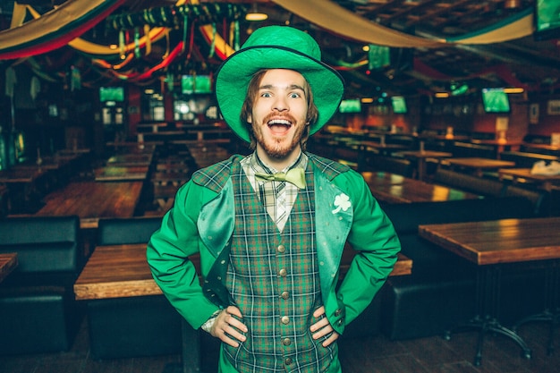 Feliz joven excitado de la mano en las caderas y sonríe a la cámara. Lleva el traje de San Patricio. Guy está solo en el pub.