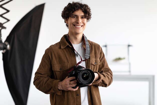 Feliz joven fotógrafo sosteniendo una cámara profesional trabajando en un moderno estudio fotográfico sonriendo a la cámara