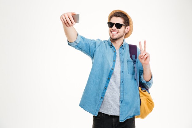 Feliz joven en gafas de sol con mochila haciendo selfie y mostrando el signo de la paz