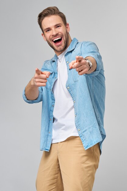 Foto feliz joven guapo en camisa de jeans apuntando a la cámara de pie