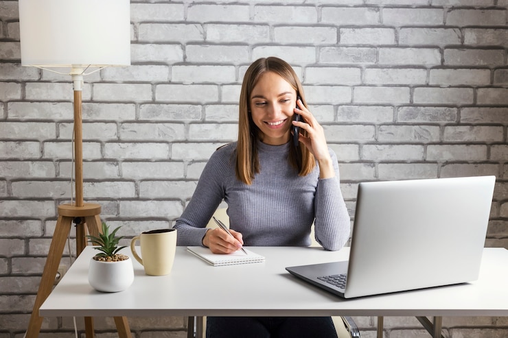 Foto feliz joven hablando por teléfono trabajando en su computadora en un escritorio blanco y pared de ladrillos grises