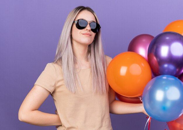 Feliz joven hermosa con gafas sosteniendo globos poniendo la mano en la cadera