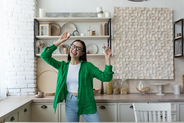 Feliz joven hermosa mujer con gafas y camisa verde bailando y cantando en casa en la cocina