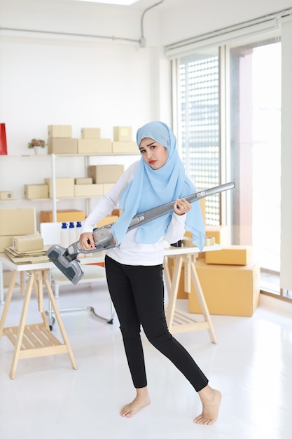 Feliz joven hermosa mujer musulmana asiática ama de casa limpiando y tocando la aspiradora como guitarra con SME y E-commerce Paquete caja de fondo. Chica de limpieza en mallas negras aspirando su habitación.