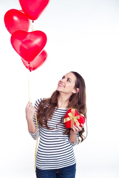 Foto feliz joven hermosa y sonriente con ropa casual con globos de helio y un regalo en forma de corazón en sus manos cumpleaños de san valentín