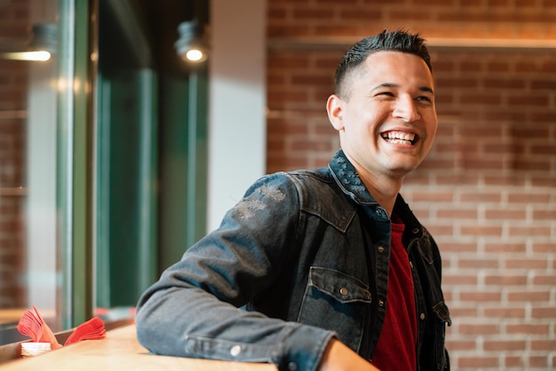 Feliz joven hispano en chaqueta de mezclilla sonriendo brillantemente mientras descansa cerca de la ventana en un café estilo loft con pared de ladrillo