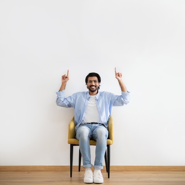 Foto feliz joven hombre del medio oriente en casual y gafas sentado en un sillón mostrando los dedos en vacío