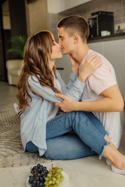 Feliz joven hombre y mujer sentados en el piso de la cocina y disfrutar de sus cabezas inclinadas el uno al otro. Pareja feliz en la elegante cocina. Foto interior de chica romántica mirando novio.