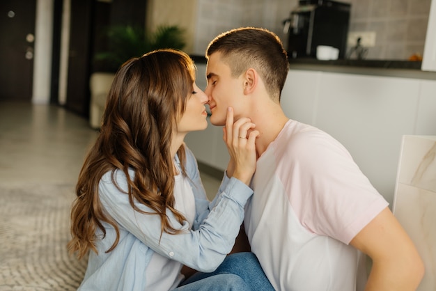 Feliz joven hombre y mujer sentados en el piso de la cocina y disfrutar de sus cabezas inclinadas el uno al otro. Pareja feliz en la elegante cocina. Foto interior de chica romántica mirando novio.