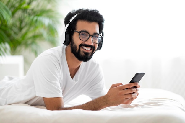 Feliz joven indio con auriculares inalámbricos descansando en la cama con un teléfono inteligente