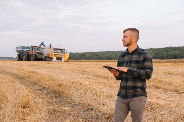 Feliz joven ingeniero agricultor con portátil de pie en el campo de trigo mientras la cosechadora trabaja en segundo plano.