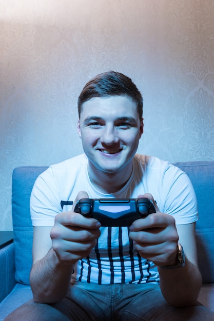 Feliz joven jugando un videojuego con una radiante sonrisa de placer sentado en el sofá frente a la televisión, vista frontal en el resplandor azul del set en casa