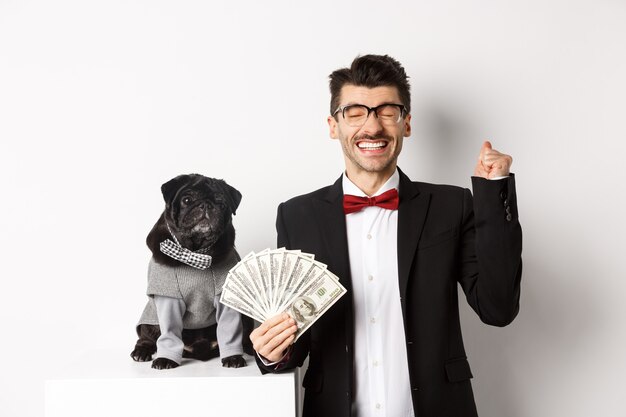 Feliz joven y lindo perro negro de pie en disfraces de fiesta, dueño de pug sosteniendo dólares de dinero y regocijándose, ganando premio, fondo blanco.
