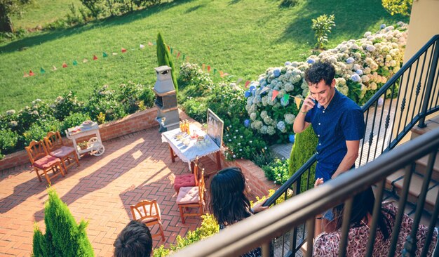 Feliz joven llamando por teléfono en las escaleras de la casa con sus amigos durante una barbacoa de verano. Concepto de estilo de vida de los jóvenes.