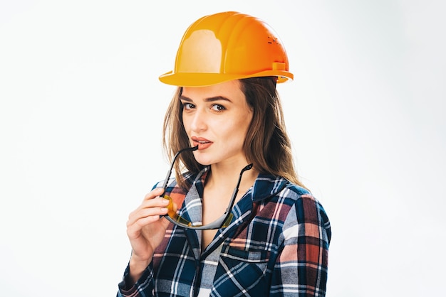 Foto feliz joven lleva casco de seguridad naranja y camisa a cuadros. mirando a cámara, gafas de seguridad amarillas cerca de los labios. fondo isoalted.