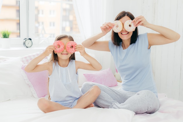 Feliz joven madre e hija se divierten juntas, sostienen deliciosas rosquillas dulces cerca de los ojos