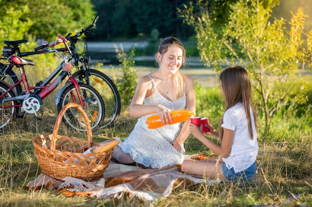 Feliz joven madre haciendo un picnic junto al río con su hija. Madre vertiendo jugo de naranja en la taza de la hija