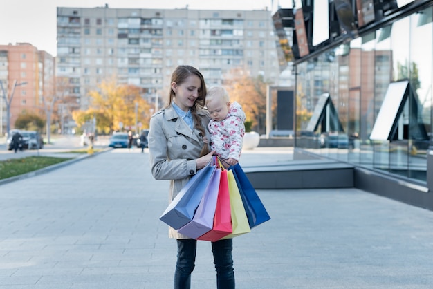 Feliz joven madre con hija pequeña en los brazos y bolsas de compras en la mano.