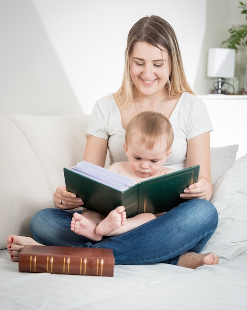 Feliz joven madre sentada en el sofá en la sala de estar y leyendo un libro a su bebé