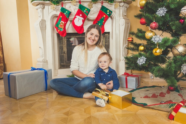 Feliz joven madre sentada con su hijo en el piso en la sala de estar junto a la chimenea y el árbol de navidad