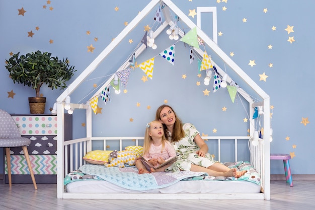 Feliz joven madre y su hija linda mirando hacia arriba mientras está sentado en la cama de la casa linda