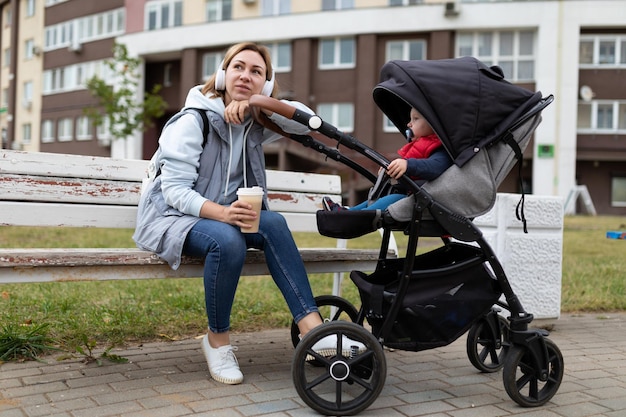 Feliz joven madre con su pequeño hijo en un cochecito camina por el parque y se relaja con café