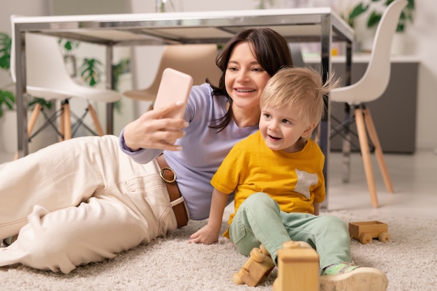 Feliz joven madre tendida en la alfombra y usando el teléfono inteligente mientras posa con el pequeño hijo para selfie