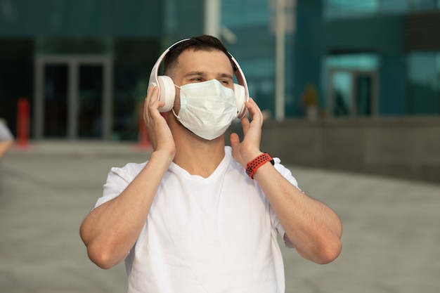 Feliz joven con mascarilla médica protectora escucha música con auriculares inalámbricos bluetooth. Coronavirus COVID-19.