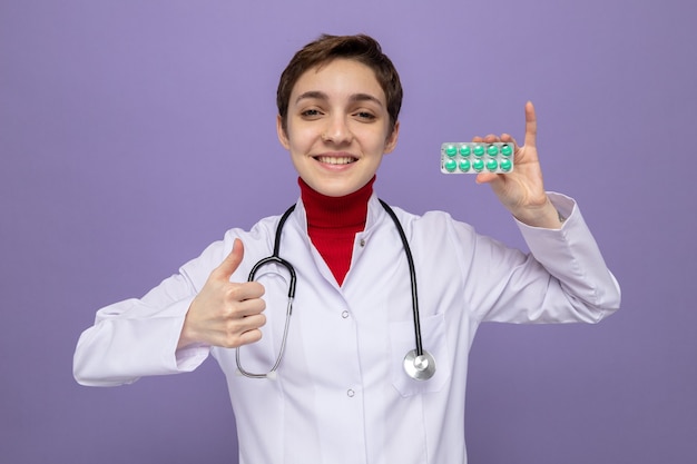 Feliz joven médico en bata blanca con estetoscopio alrededor del cuello sosteniendo blister con pastillas sonriendo alegremente mostrando los pulgares para arriba de pie sobre la pared púrpura