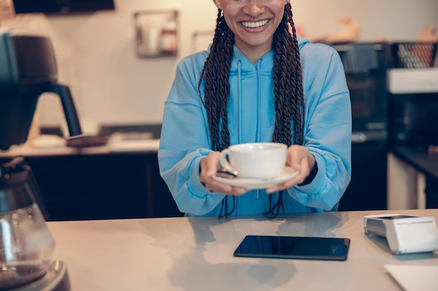 Foto feliz joven mestiza mujer barista con trenzas dando y colgando una taza de café a la cámara