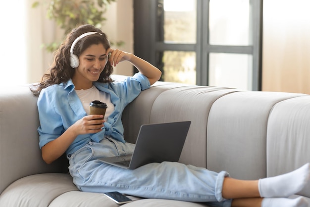 Feliz joven morena con auriculares teniendo videoconferencia en línea en una laptop tomando café en