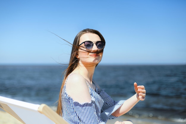 Feliz joven morena relajándose en una tumbona de madera en la playa del océano mientras mira la cámara, sonríe y usa gafas de sol de moda. El concepto de disfrutar de las vacaciones