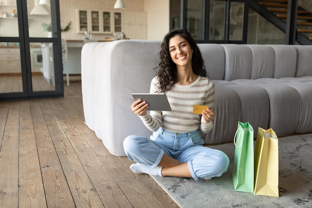 Feliz joven morena con tableta digital y bolsas de regalo usando tarjeta de crédito mientras compra en línea