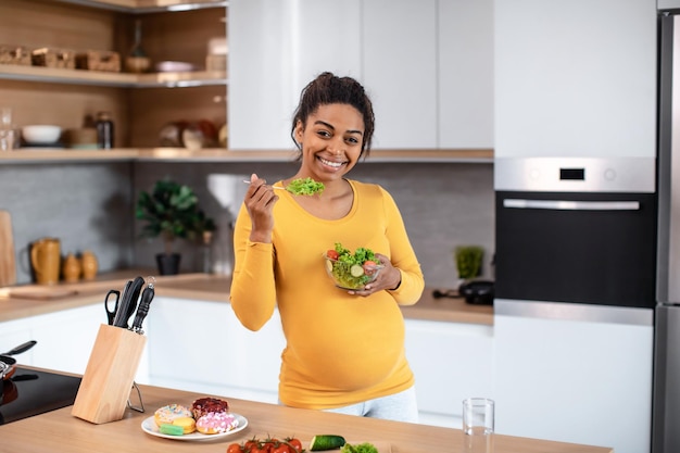 Feliz joven mujer afroamericana embarazada sostener plato con ensalada en la mesa con verduras y donas