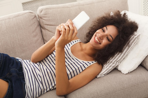 Feliz joven mujer afroamericana enviando mensajes en el teléfono inteligente en casa, cómodamente acostado en el sofá beige, espacio de copia