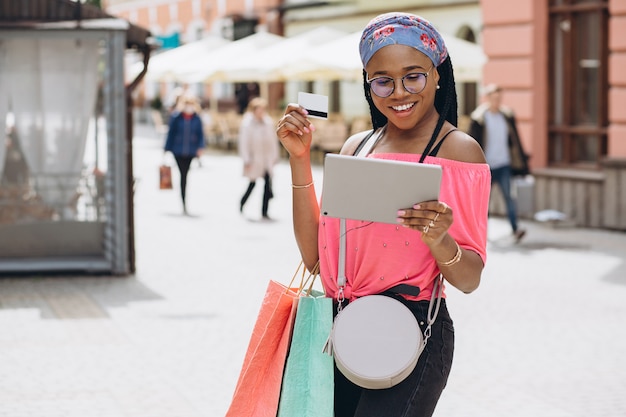 Feliz joven mujer afroamericana hacer compras en la tableta con tarjeta de crédito y sosteniendo bolsas de compras en la calle